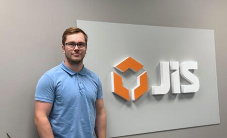 Nuori mies seisoo vaaleanharmaata seinää vasten sinisessä kauluspaidassa. Hänen vieressään on suuri valkoinen kyltti, jossa on oranssi-logo ja teksti JIS.